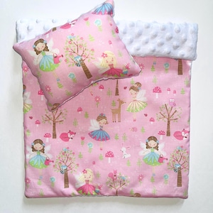 May include: A pink baby blanket and pillow set featuring a fairy and woodland creature pattern. The blanket has a white, textured border. The pillow and blanket are decorated with fairies, trees, mushrooms, and animals.
