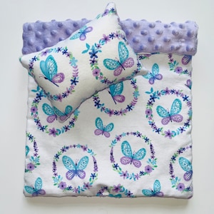 May include: A purple and white doll bed with a floral print featuring butterflies. The bed has a matching pillow with a white background and a purple and blue butterfly design.