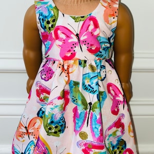 K&ouml;nnte beinhalten: Ein &auml;rmelloses Kleid mit hellrosa Hintergrund und einem lebendigen, mehrfarbigen Schmetterlingsdruck. Das Muster zeigt Schmetterlinge in Pink, leuchtendem Blau, Orange und Gr&uuml;n, mit einer gerafften Taille und einem Rundhalsausschnitt.
