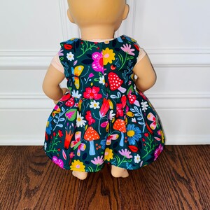 May include: A green dress with a floral and mushroom print for a doll. The dress has a gathered skirt and a sleeveless bodice.