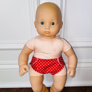 May include: A bald baby doll with blue eyes wearing a pink short-sleeved shirt with a pink star and red and white polka dot shorts.