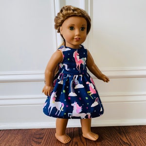 May include: A doll wearing a navy blue dress with a unicorn print. The dress features pink, blue and yellow unicorns, rainbows and stars.