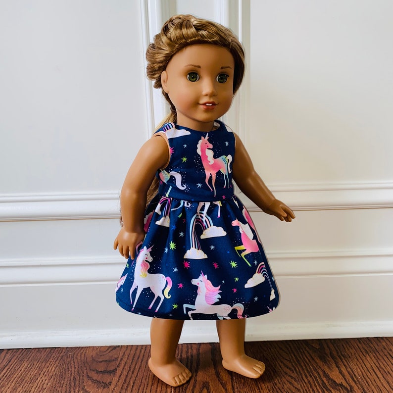 May include: A doll wearing a navy blue dress with a unicorn print. The dress features pink, blue and yellow unicorns, rainbows and stars.