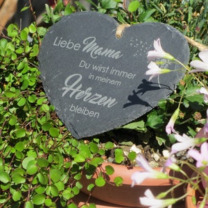 K&ouml;nnte beinhalten: Eine herzf&ouml;rmige Schiefertafel mit dem Text "Liebe Mama Du wirst immer in meinem Herzen bleiben" in wei&szlig;er Schrift. Die Tafel ist von gr&uuml;nen Pflanzen und rosa Bl&uuml;ten umgeben.