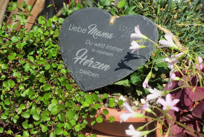 K&ouml;nnte beinhalten: Eine herzf&ouml;rmige Schiefertafel mit dem Text "Liebe Mama Du wirst immer in meinem Herzen bleiben" in wei&szlig;er Schrift. Die Tafel ist von gr&uuml;nen Pflanzen und rosa Bl&uuml;ten umgeben.