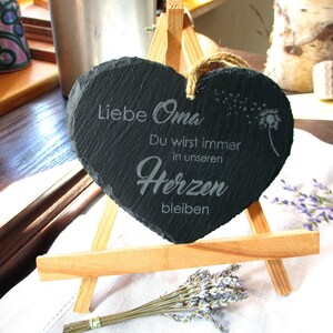 K&ouml;nnte beinhalten: Eine schwarze herzf&ouml;rmige Schiefertafel mit dem Text "Liebe Oma Du wirst immer in unseren Herzen bleiben" (Liebe Oma Du wirst immer in unseren Herzen bleiben) und einem L&ouml;wenzahnmuster. Die Tafel steht auf einem kleinen Holzst&auml;nder.