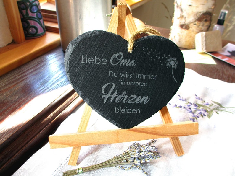 K&ouml;nnte beinhalten: Eine schwarze herzf&ouml;rmige Schiefertafel mit dem Text "Liebe Oma Du wirst immer in unseren Herzen bleiben" (Liebe Oma Du wirst immer in unseren Herzen bleiben) und einem L&ouml;wenzahnmuster. Die Tafel steht auf einem kleinen Holzst&auml;nder.