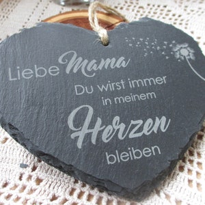 K&ouml;nnte beinhalten: Eine schwarze herzf&ouml;rmige Schiefertafel mit dem Text "Liebe Mama Du wirst immer in meinem Herzen bleiben" und einem L&ouml;wenzahn mit Samen, die im Wind fliegen.