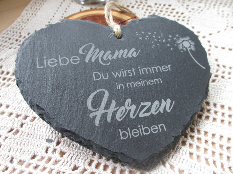 K&ouml;nnte beinhalten: Eine schwarze herzf&ouml;rmige Schiefertafel mit dem Text "Liebe Mama Du wirst immer in meinem Herzen bleiben" und einem L&ouml;wenzahn mit Samen, die im Wind fliegen.