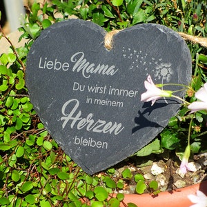 K&ouml;nnte beinhalten: Eine herzf&ouml;rmige Schiefertafel mit dem Text "Liebe Mama Du wirst immer in meinem Herzen bleiben" (Liebe Mama Du wirst immer in meinem Herzen bleiben). Die Tafel ist von gr&uuml;nen Pflanzen und rosa Bl&uuml;ten umgeben.