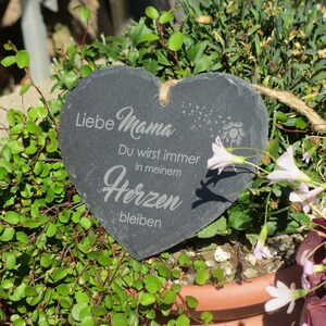 K&ouml;nnte beinhalten: Eine schwarze herzf&ouml;rmige Schiefertafel mit dem Text "Liebe Mama Du wirst immer in meinem Herzen bleiben" in wei&szlig; graviert. Die Tafel h&auml;ngt an einer Bindfadenkordel und ist von gr&uuml;nen Pflanzen und rosa Bl&uuml;ten umgeben.
