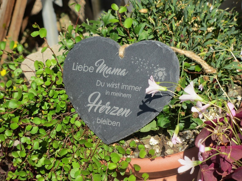 K&ouml;nnte beinhalten: Eine schwarze herzf&ouml;rmige Schiefertafel mit dem Text "Liebe Mama Du wirst immer in meinem Herzen bleiben" in wei&szlig; graviert. Die Tafel h&auml;ngt an einer Bindfadenkordel und ist von gr&uuml;nen Pflanzen und rosa Bl&uuml;ten umgeben.