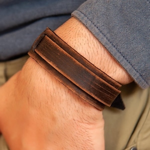 May include: A brown leather bracelet with a simple design. The bracelet is worn on a person's wrist.