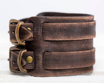 May include: A brown leather bracelet with two brass buckles. The bracelet is made of multiple layers of leather, giving it a rugged look.