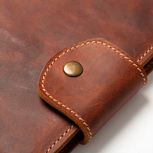May include: Close-up of a brown leather notebook with a flap closure and a brass button. The notebook has white stitching around the edges.