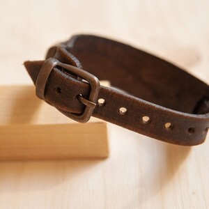 May include: A brown leather bracelet with a silver buckle and multiple holes for adjusting the size.