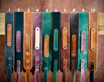 May include: Ten leather camera straps with personalized name tags. The straps are in various colors, including brown, green, purple, and black. The name tags are in a cream color with black lettering.