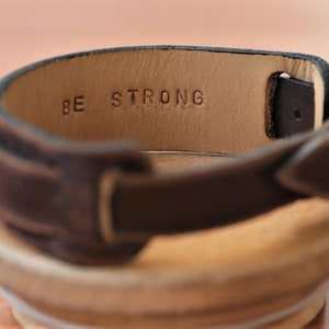 May include: A brown leather watch strap with a silver buckle. The strap has the words "BE STRONG" embossed on the inside.