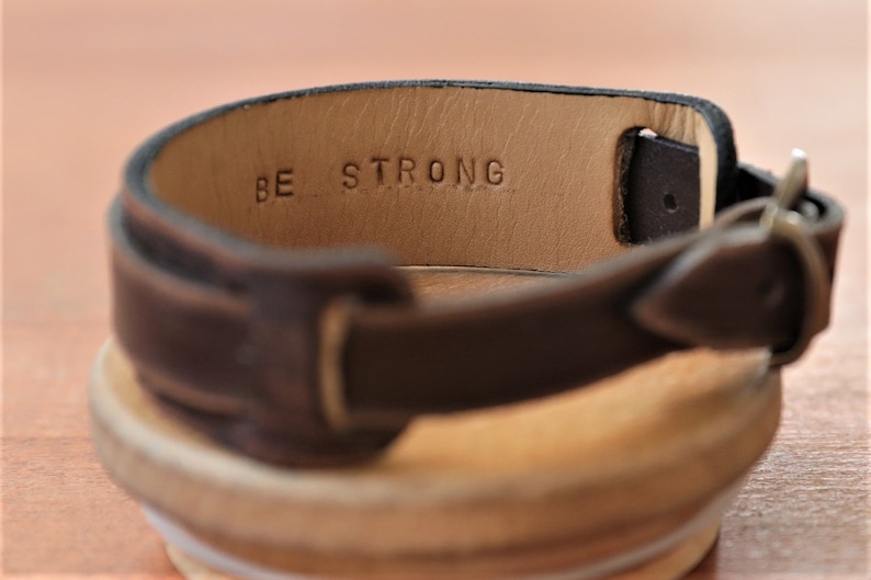 May include: A brown leather watch strap with a silver buckle. The strap has the words "BE STRONG" embossed on the inside.