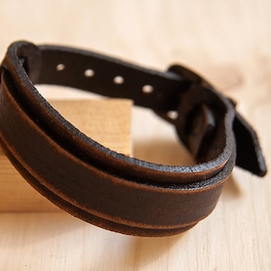 May include: A brown leather bracelet with a buckle closure. The bracelet is made of two layers of leather and has a distressed look.