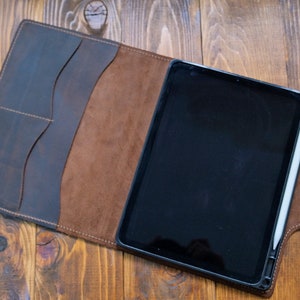 May include: A brown leather folio case with a flap that opens to reveal a black tablet and a white stylus. The case has multiple pockets and a brown suede lining.