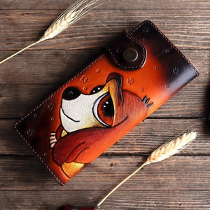 May include: A brown leather wallet featuring a cartoon animal illustration. The animal has large eyes and is painted in shades of orange, brown, and white.