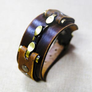 May include: A brown leather bracelet with gold metal accents. The bracelet has a simple design and is made of high-quality materials.