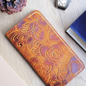 May include: A brown leather wallet with a floral embossed design. The wallet has a stitched edge and is lying on a wooden surface.