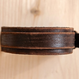 May include: A brown leather bracelet with a simple design. The bracelet is made of two layers of leather, with the top layer being slightly wider than the bottom layer.