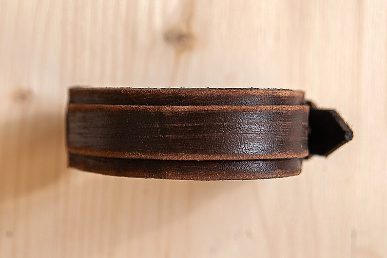 May include: A brown leather bracelet with a simple design. The bracelet is made of two layers of leather, with the top layer being slightly wider than the bottom layer.