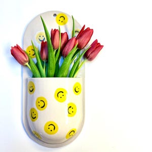 Puede incluir: Un jarrón de pared de cerámica blanca con caras sonrientes amarillas, que contiene tulipanes rojos y hojas verdes. El jarrón está diseñado para montaje en pared y tiene forma redondeada.