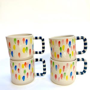 Può includere: Quattro tazze in ceramica bianca con manici a righe blu e bianche. Ogni tazza è decorata con un motivo a coriandoli colorati.