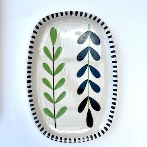 May include: An oval ceramic platter with a cream-colored base and a black dashed border. The center features two vertical botanical illustrations: one stem with light green leaves and another stem with dark navy blue leaves painted on the surface.