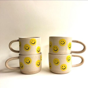 May include: Set of four ceramic mugs with yellow smiley faces painted on a beige background. The mugs have a handle and are stacked in pairs.