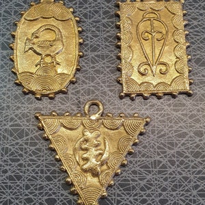 May include: Three gold-coloured pendants with detailed patterns. An oval pendant features a bird design, a rectangular one has a stylized symbol, and a triangular pendant displays a unique emblem. Each has a loop for hanging and a textured border.