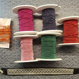 Op de afbeelding: Vijf spoelen elastisch koord in magenta, donkerpaars, rood, lichtroze en groen, plus een plat oranje koord op een witte kaart. Daaronder ligt een donkerbruine leren armband met een zilverkleurig vlechtpatroon en een kleine witte knoopsluiting.