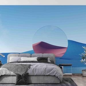 May include: A bedroom featuring a grey upholstered bed with a grey textured throw blanket and pillows, positioned against a wall mural depicting a blue desert landscape with a large circular cutout revealing a pink sand dune under a clear blue sky.