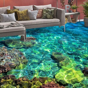 May include: A living room with a unique floor design featuring a vibrant, realistic depiction of clear, turquoise water over a rocky seabed. A gray sofa with various throw pillows sits on the floor, creating a seamless transition between the furniture and the aquatic scene.
