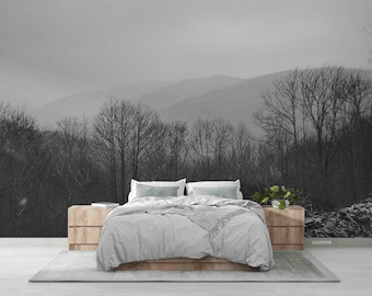 May include: A white bed with a gray duvet cover and two white pillows sits in front of a black and white photo of a mountain range with trees in the foreground.