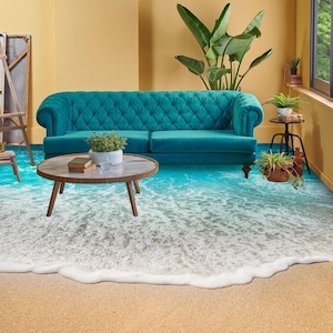 May include: A teal blue tufted sofa sits in a room with a beach-themed floor. The floor is a 3D optical illusion of a sandy beach with blue water and white waves. A wooden coffee table with books sits in front of the sofa.