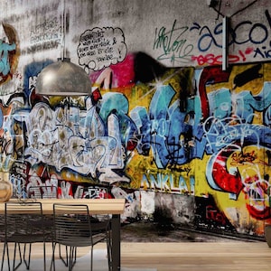 May include: A colorful graffiti wall mural with a dining room table and chairs in the foreground. The wall is covered in vibrant colors, including red, yellow, blue, green, and purple. The graffiti is in a variety of styles, including bubble letters, block letters, and abstract designs. The dining room table is made of wood and has four chairs. The chairs are black metal with a mesh back.