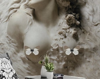 May include: A white plaster sculpture of a woman's bust with a floral crown and butterflies on her chest. The sculpture is set against a textured wall and a gray blanket is draped over a white couch in the foreground.