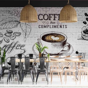Op de afbeelding: Een café-interieur met een koffiethema muurschildering op een witte bakstenen muur. De muurschildering toont koffiebonen, bladeren en een kop koffie met de tekst "Coffee than Compliments". Rotan lampen hangen boven tafels en stoelen.