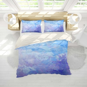 Peut inclure: Une housse de couette et deux taies d'oreiller assorties avec un motif abstrait bleu et violet. La literie est sur un cadre de lit blanc avec un tapis blanc moelleux au sol.