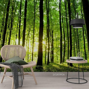 Pode incluir: Uma vibrante cena de floresta com árvores altas e luz solar a atravessar as folhas. Uma cadeira de cor clara com uma almofada verde e um cobertor cinzento escuro está em primeiro plano. Uma pequena mesa lateral com livros e uma lâmpada preta completam a cena.