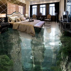 Op de afbeelding: Een slaapkamer met een groot bed, twee stoelen en een bijzettafel. De vloer is bedekt met een 3D-geprinte vloerbedekking die eruitziet als een bergketen met een rivier die erdoorheen stroomt. Er vliegen vogels over de bergen.