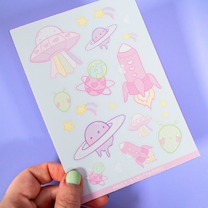 May include: A sheet of stickers featuring a variety of space-themed illustrations, including planets, rockets, aliens, and UFOs. The stickers are in pastel colors, including pink, purple, green, and blue. The sheet also includes the text "Nancy Illustrations".