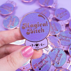 Könnte beinhalten: Lila Emaille-Anstecknadeln mit Glitzer-Finish und roségoldenen Akzenten. Jede Anstecknadel zeigt ein Kristallkugel-Design mit dem Schriftzug "Magical Bitch" in einer Retro-Schriftart. Die Anstecknadeln haben eine niedliche, cartoonartige Ästhetik.