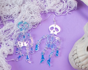 May include: A pair of iridescent skeleton earrings with a skull at the top. The earrings are hanging from silver hooks and are on a purple background.