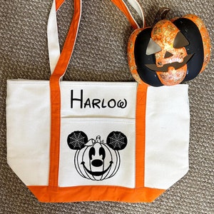 K&ouml;nnte beinhalten: Wei&szlig;e Canvas-Tragetasche mit orangefarbenem Besatz. Die Tasche hat ein schwarz-wei&szlig;es Bild von Mickey Mouse, der als K&uuml;rbis verkleidet ist, mit einem Spinnennetz an den Ohren. Die Tasche tr&auml;gt auch den Namen "HARLOW" auf sich.